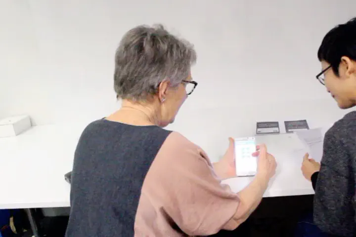 An older adult user sits on the left of the image, holding a smartphone with the Prospecfit app open in a think-aloud testing session. Researcher sits on the right, listening to what the user is saying.